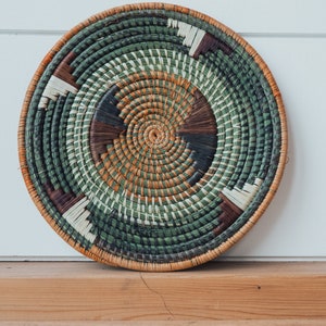 May include: A round woven wall hanging with a green, brown, and white pattern. The hanging is made of natural materials and has a circular shape.