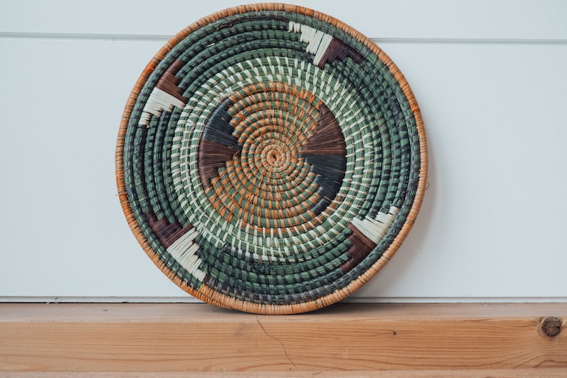 May include: A round woven wall hanging with a green, brown, and white pattern. The hanging is made of natural materials and has a circular shape.