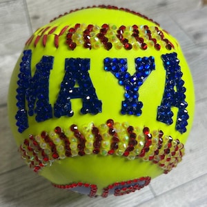 May include: A bright yellow softball with the name "MAYA" in blue rhinestones. Red and yellow rhinestone accents adorn the seams. The stitching is red.