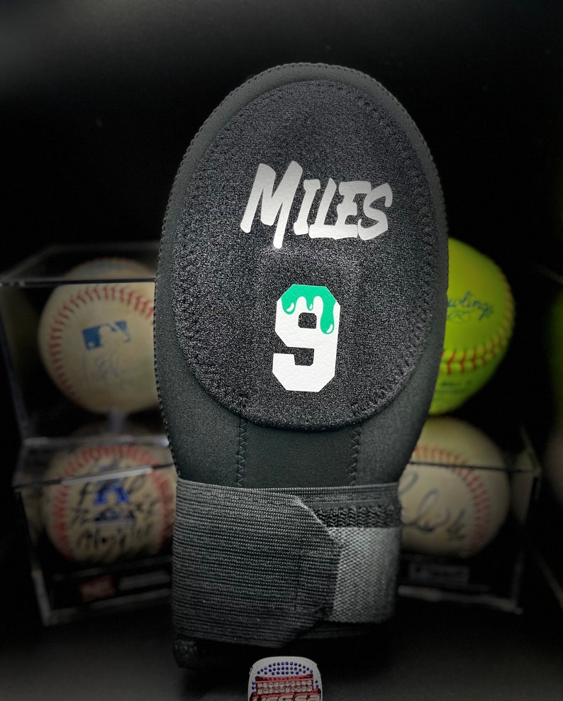 Puede incluir: Guante de b&eacute;isbol negro con el nombre "Miles" en escritura blanca y el n&uacute;mero "9" con un dise&ntilde;o de goteo verde. El guante tiene una superficie texturizada y una correa el&aacute;stica negra para la mu&ntilde;eca. Se ven pelotas de b&eacute;isbol y s&oacute;ftbol.