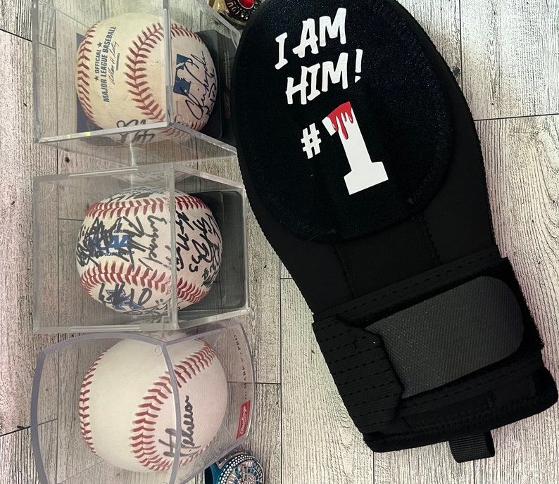 Puede incluir: Una mu&ntilde;equera negra con el texto "I AM HIM! #1" en blanco y rojo. Tambi&eacute;n se ven tres pelotas de b&eacute;isbol en vitrinas transparentes. Las pelotas son blancas con costuras rojas y firmas.