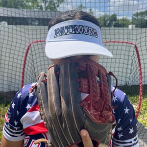 May include: A person wearing a white visor with the word "PATRIOTS" in black and red letters. The visor has a red, white, and blue American flag design. The person is holding a brown leather baseball glove in front of their face. The person is wearing a red, white, and blue American flag patterned shirt.