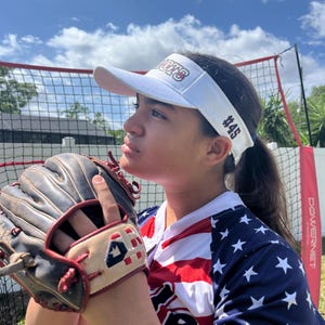 May include: A young person wearing a white visor with the text "Patriots #45" in maroon, a white and red, white and blue stars baseball jersey, and a brown leather baseball glove.