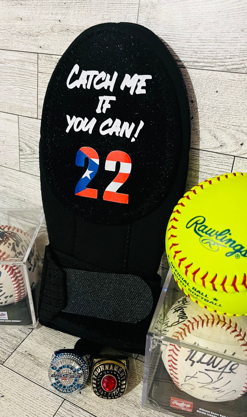 Puede incluir: Guante de b&eacute;isbol negro con el texto "CATCH ME IF YOU CAN!" y el n&uacute;mero 22 en rojo, blanco y azul. Tambi&eacute;n se ven una pelota de s&oacute;ftbol amarilla y dos pelotas de b&eacute;isbol, junto con dos anillos de campeonato.