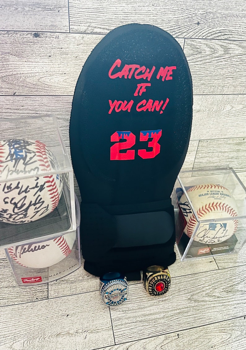 Puede incluir: Un guante de b&eacute;isbol negro con el texto "CATCH ME IF YOU CAN!" y el n&uacute;mero 23 en rojo. Dos pelotas de b&eacute;isbol en estuches transparentes, una firmada. Tambi&eacute;n se ven dos anillos de campeonato.