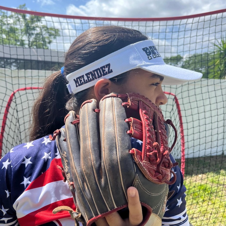 May include: A young person wearing a white visor with the text "MELENDEZ" and "PR" on it. They are holding a brown and red baseball glove. The person is wearing a red, white, and blue shirt with stars.