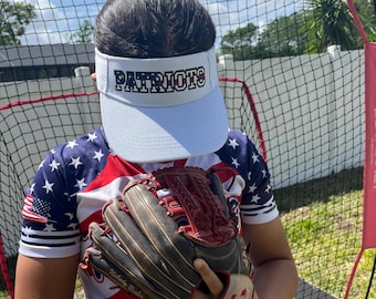 May include: A person wearing a white visor with the word "PATRIOTS" in black lettering, a red, white, and blue American flag shirt, and a brown baseball glove.