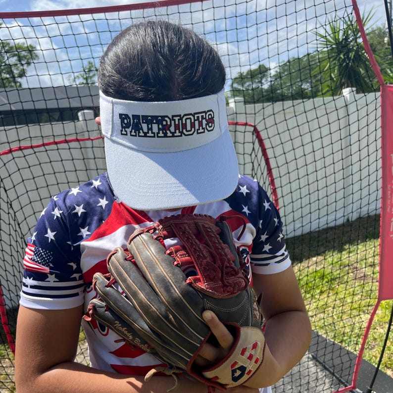 May include: A person wearing a white visor with the word "PATRIOTS" in black lettering, a red, white, and blue American flag shirt, and a brown baseball glove.