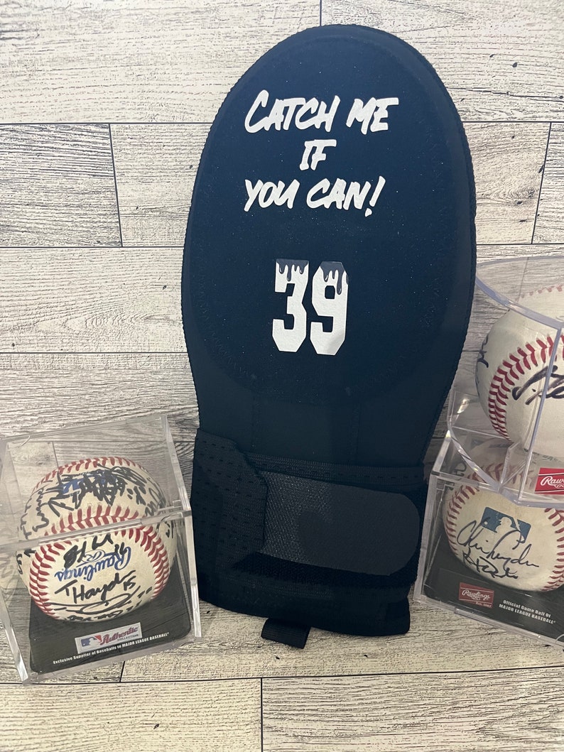 Puede incluir: Un guante de receptor de b&eacute;isbol negro con el texto "Catch Me If You Can!" y el n&uacute;mero 39 en blanco. Tambi&eacute;n se ven dos pelotas de b&eacute;isbol en estuches transparentes, cada una con firmas.