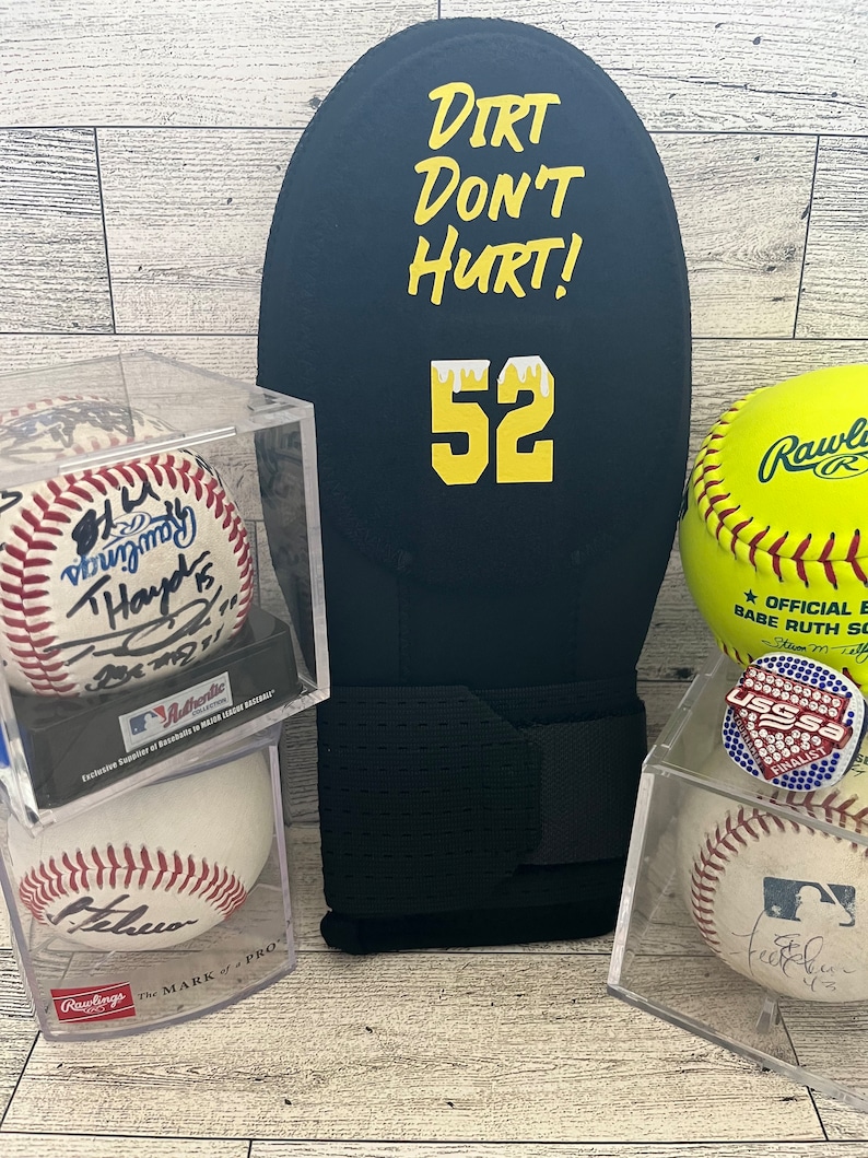 Puede incluir: Un guante de b&eacute;isbol negro con el texto "Dirt Don't Hurt!" y el n&uacute;mero 52 en amarillo. Se exhiben varias pelotas de b&eacute;isbol y una de softbol, algunas en vitrinas transparentes. Una pelota de b&eacute;isbol est&aacute; firmada.