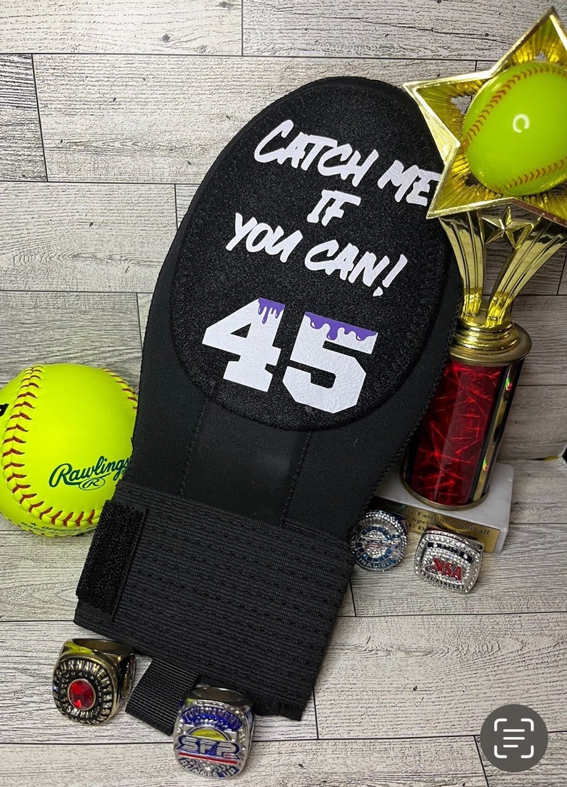 Puede incluir: Mu&ntilde;equera de softbol negra con el texto "CATCH ME IF YOU CAN!" y el n&uacute;mero 45 en blanco. Una pelota de softbol amarilla, un trofeo y varios anillos de campeonato tambi&eacute;n est&aacute;n en la imagen.