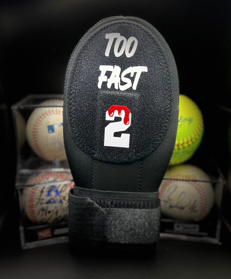 Puede incluir: Empu&ntilde;adura de bate de b&eacute;isbol negra con el texto "TOO FAST 2" en blanco. El n&uacute;mero 2 tiene un dise&ntilde;o de goteo rojo. Se ven pelotas de b&eacute;isbol y una de s&oacute;ftbol.