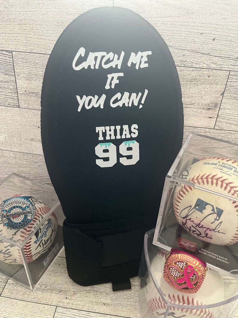 Puede incluir: Art&iacute;culo negro en forma de bal&oacute;n de f&uacute;tbol con el texto "CATCH ME IF YOU CAN!" y "THIAS 99" en blanco. Recuerdos de b&eacute;isbol, incluyendo pelotas firmadas y anillos de campeonato, se exhiben en estuches transparentes.