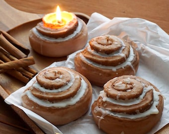 Könnte beinhalten: Ein Holztablett mit vier Zimtschnecken-Kerzen mit weißem Zuckerguss. Eine Kerze ist angezündet, mit einer gelben Flamme. Zimtstangen und eine goldene Gabel befinden sich ebenfalls auf dem Tablett. Die Kerzen liegen auf weißem Papier.