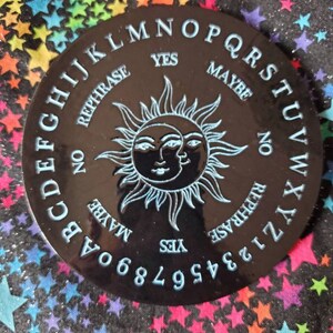 May include: A black circular ouija board with a sun and moon design. The board has letters, numbers, and the words "yes", "no", "maybe", and "rephrase".
