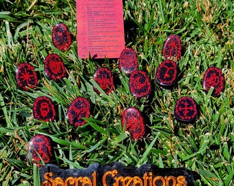 May include: A set of thirteen oval-shaped black stones with red glitter and hand-painted red symbols, accompanied by a rectangular red information card titled Witch Runes and a black plaque with the orange text Sacral Creations.
