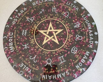 May include: A black circular resin art piece with a gold pentagram in the center. The piece is decorated with rose petals and features the names of the eight pagan holidays in a circle around the edge, along with the astrological signs and dates.