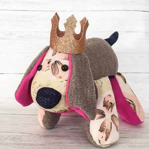 May include: A plush toy dog wearing a gold glitter crown. The dog is made of fabric with a floral pattern and has pink accents.