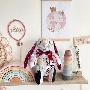 May include: A colourful handmade stuffed bunny with a rainbow pattern and the word "Mummy" written on its chest. The bunny is sitting on a wooden shelf with a rainbow stacking toy, a pink balloon with the word "play" on it, and a framed print of a pink earth with a crown on top.