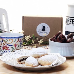May include: A white ceramic cup with a colorful floral pattern sits on a white lace doily. The cup is filled with coffee and there are two cookies on the doily. The cookies are covered in powdered sugar and have a brown filling. There is a white ceramic bowl filled with dates in the background. The bowl is sitting on a wooden table. The text "OFFEE" and "THANK YOU" are visible on a black and white sign in the background.