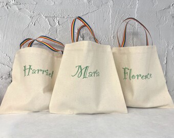 May include: Three white canvas tote bags with rainbow striped handles. The bags are personalized with the names "Harriet", "Maria", and "Florence" in green embroidery.