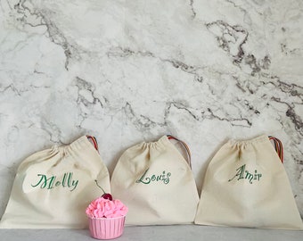 May include: Three white cotton drawstring bags with green embroidered names: Molly, Louis, and Almir. A pink cupcake with a cherry on top sits in front of the bags.