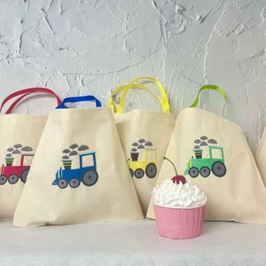 May include: Four white canvas tote bags with colorful handles and embroidered train designs. Each bag features a different colored train with black smoke and gray clouds. A pink cupcake with a red cherry on top sits in front of the bags.