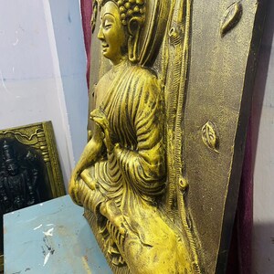 May include: A gold-colored, three-dimensional wall sculpture of a seated Buddha. The sculpture is set against a textured, dark brown background.