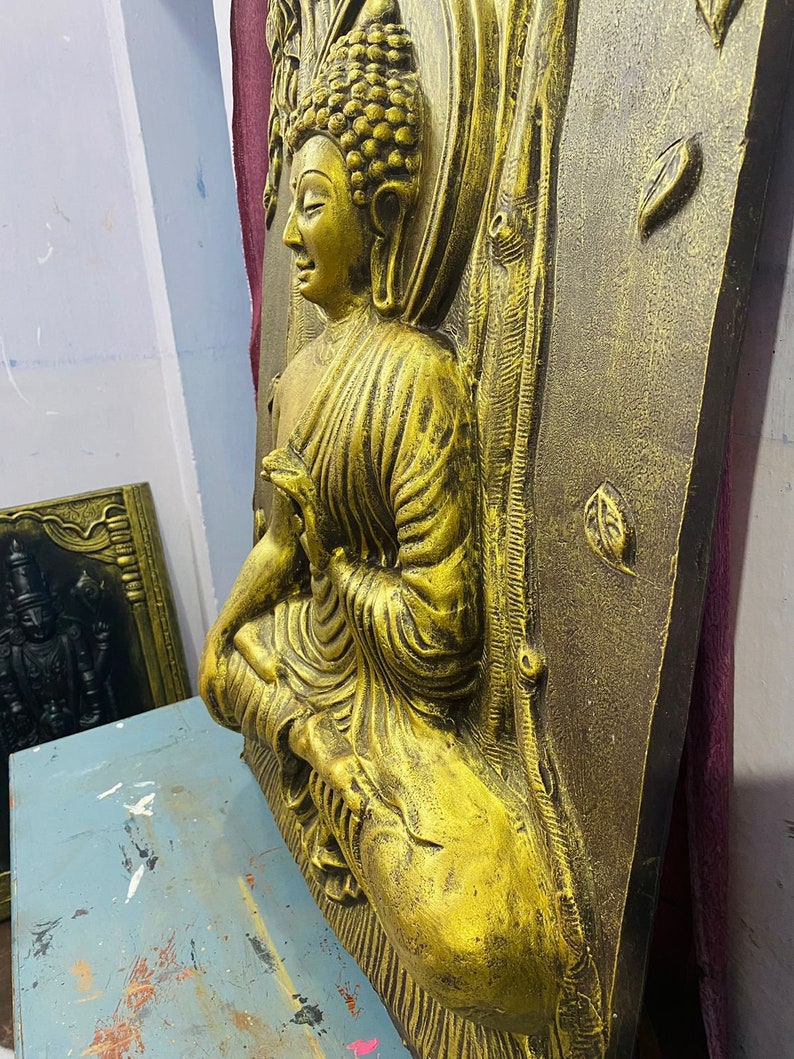 May include: A gold-colored, three-dimensional wall sculpture of a seated Buddha. The sculpture is set against a textured, dark brown background.