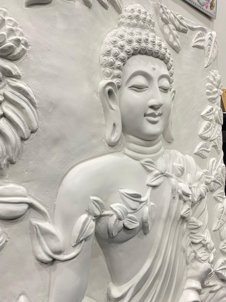 May include: White plaster wall sculpture of a Buddha with a floral design. The Buddha is depicted in a seated meditation pose with a serene expression. The sculpture is detailed with intricate carvings of leaves and flowers.