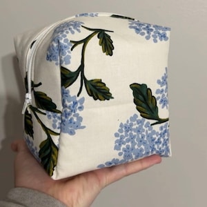 Puede incluir: Un neceser blanco en forma de cubo con un estampado floral de hortensias azules y hojas verdes. La bolsa tiene una cremallera blanca y se sostiene en una mano.