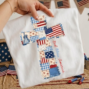 Puede incluir: Tela blanca con un diseño de cruz hecho con piezas de tela de patchwork. La cruz presenta la bandera estadounidense, patrones florales y la Estatua de la Libertad. Las palabras "God Bless America" están bordadas en rojo.