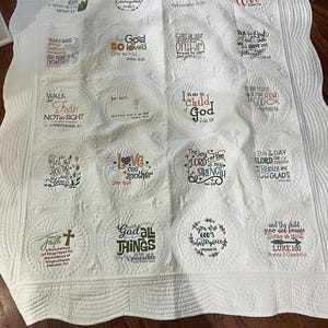 May include: A white quilted blanket with various inspirational quotes embroidered in different colors. The quotes are centered in circles and include verses from the Bible. The blanket is a great way to add a touch of faith and inspiration to any room.