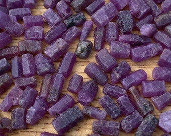 May include: A collection of purple rough gemstones, likely amethyst or sapphire, in various sizes and shapes. The stones are raw and unpolished, with a natural, irregular appearance.
