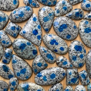 May include: A collection of polished blue and gray speckled stones with a variety of shapes and sizes. The stones are arranged on a wooden surface.