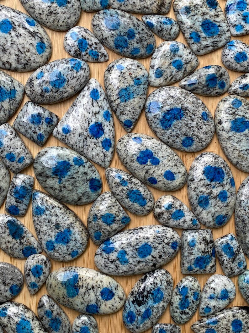 May include: A collection of polished blue and gray speckled stones with a variety of shapes and sizes. The stones are arranged on a wooden surface.