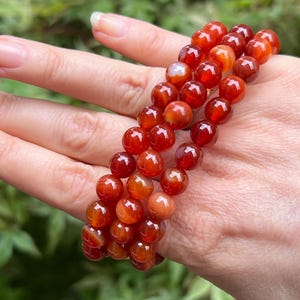 May include: A multi-strand bracelet composed of polished, spherical carnelian beads in varying shades of deep orange, burnt red, and translucent amber. The beads are strung together in a circular formation, showcasing natural color variations.