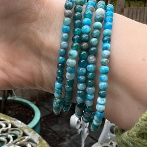May include: A stack of five blue and green gemstone bracelets. The bracelets are made of small, round beads and are stacked on a person's wrist.