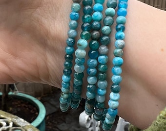 May include: A stack of five blue and green gemstone bracelets. The bracelets are made of small, round beads and are stacked on a person's wrist.