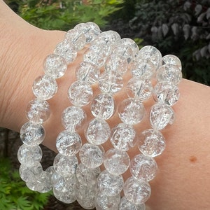 May include: Clear, crackle quartz beaded bracelets. The bracelets are made of round, transparent beads with internal fractures, strung together. The bracelets are worn on a wrist, with a blurred background of greenery and a building.