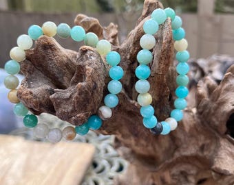May include: Two beaded bracelets with light blue, green, and white beads. The bracelets are stacked on top of each other and are resting on a piece of driftwood.