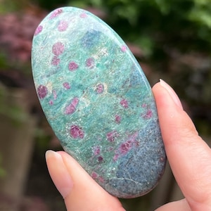 May include: A green and pink oval-shaped polished stone with red and white inclusions. The stone is smooth and has a speckled appearance.