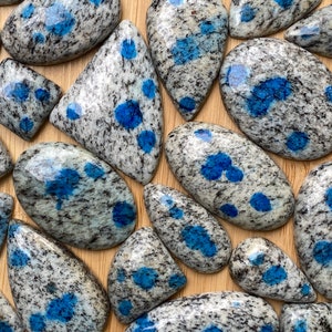 May include: A collection of polished blue and white speckled cabochons, also known as blue kyanite granite, on a wooden surface.