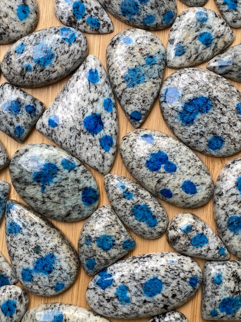 May include: A collection of polished blue and white speckled cabochons, also known as blue kyanite granite, on a wooden surface.
