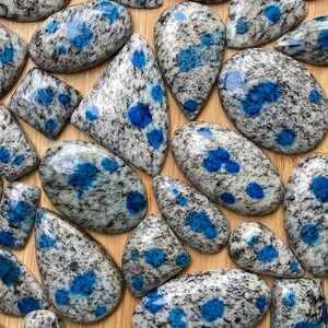 May include: A collection of polished blue and gray speckled stone cabochons. The stones are various shapes and sizes, with a smooth, polished finish.