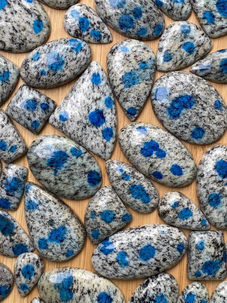 May include: A collection of polished blue and gray speckled stone cabochons. The stones are various shapes and sizes, with a smooth, polished finish.