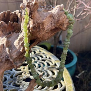 Op de afbeelding: Een ketting van lichtgroene edelstenen met onregelmatig gevormde kralen. De ketting wordt getoond op een stuk drijfhout, met een metalen sluiting. De achtergrond omvat een decoratieve metalen tafel en een blauwe bloempot.