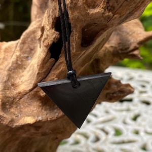 May include: A black triangular pendant necklace. The pendant is suspended from a black cord. The necklace is displayed against a natural wood background. The pendant is a simple geometric shape.