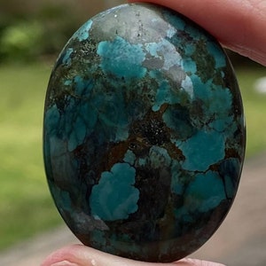 May include: A smooth, oval-shaped turquoise cabochon with a dark green and blue marbled pattern. The stone has a rich, earthy colour and a polished finish.