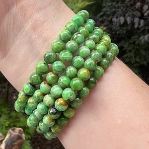 Puede incluir: Varios brazaletes de cuentas verdes. Las cuentas son redondas y presentan un patrón moteado con tonos verdes y marrones. Los brazaletes se llevan en una muñeca, mostrando el color y la textura de la joyería.